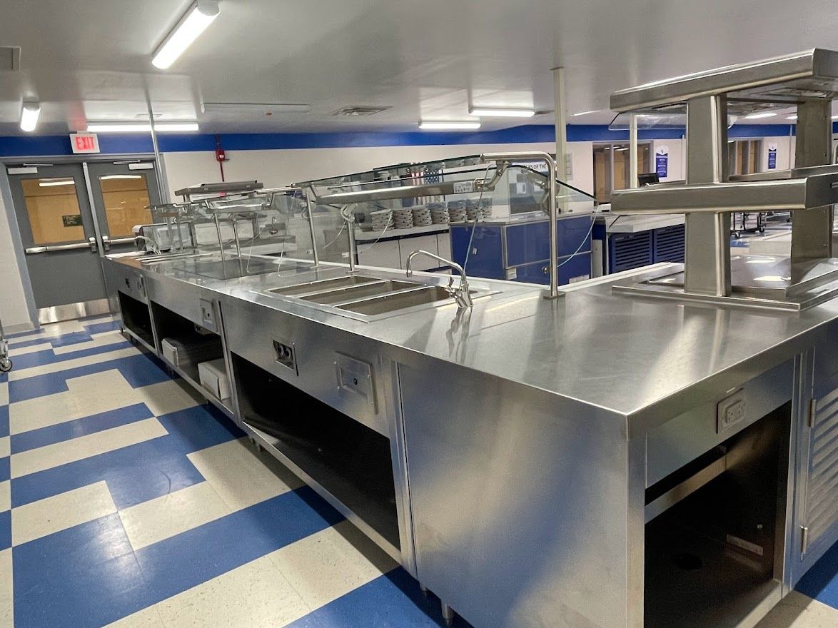 A long stainless steel cafeteria serving counter with glass sneeze guards set against a blue and white tiled floor.