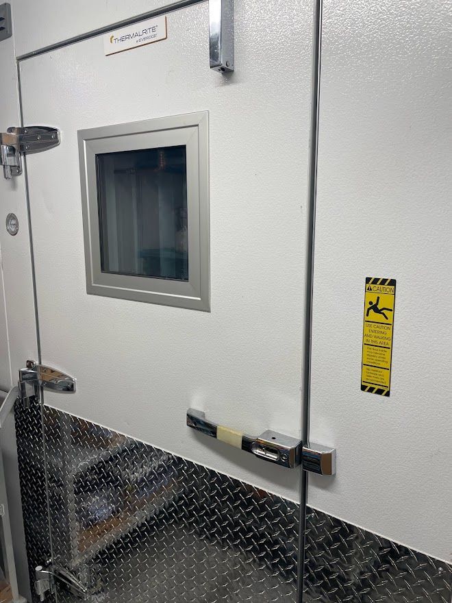 White walk-in cooler door with a window, diamond-plate kickplate, metal handle, and yellow warning sign.