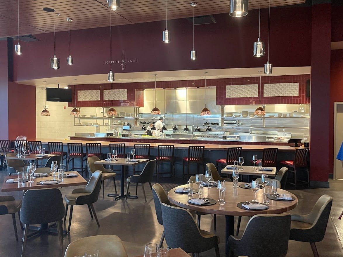 An elegant dining area with dark red walls, a stainless steel kitchen counter, pendant lighting, and wooden tables.