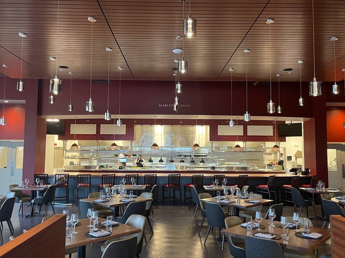 A restaurant interior with tables, a long bar, and numerous pendant lights hanging from a wood-paneled ceiling.