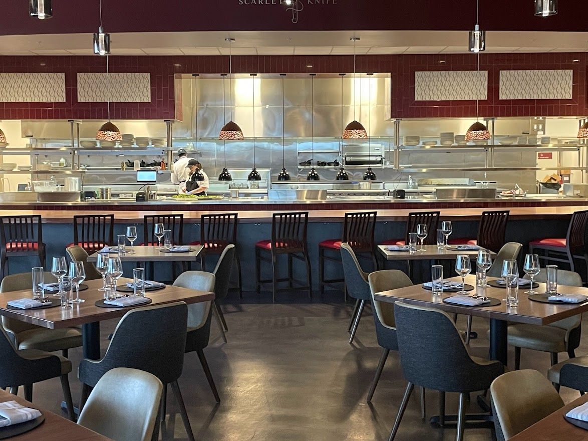 A modern restaurant interior featuring wooden tables, dark upholstered chairs, and an open kitchen with a metal counter.