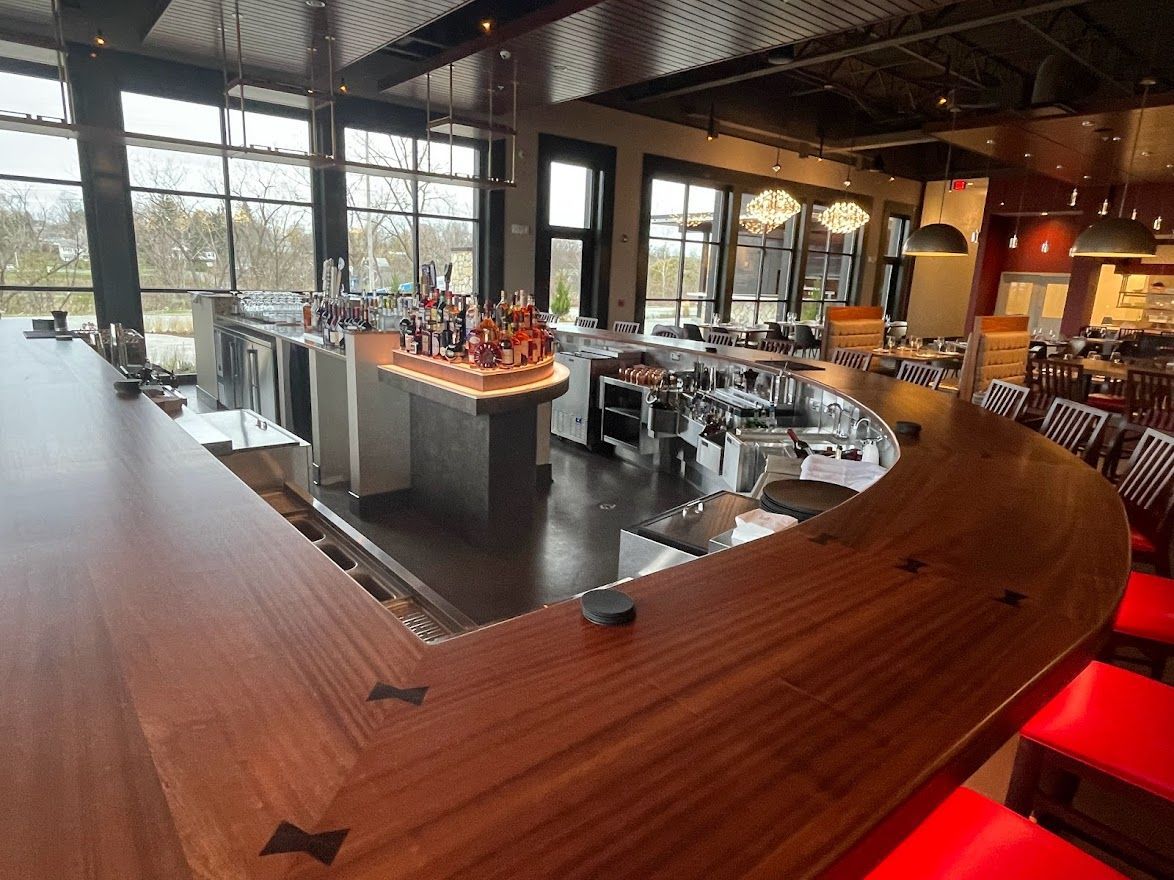 A curved wooden bar with bow-tie joinery, overlooking a modern restaurant dining room with large windows and red chairs.