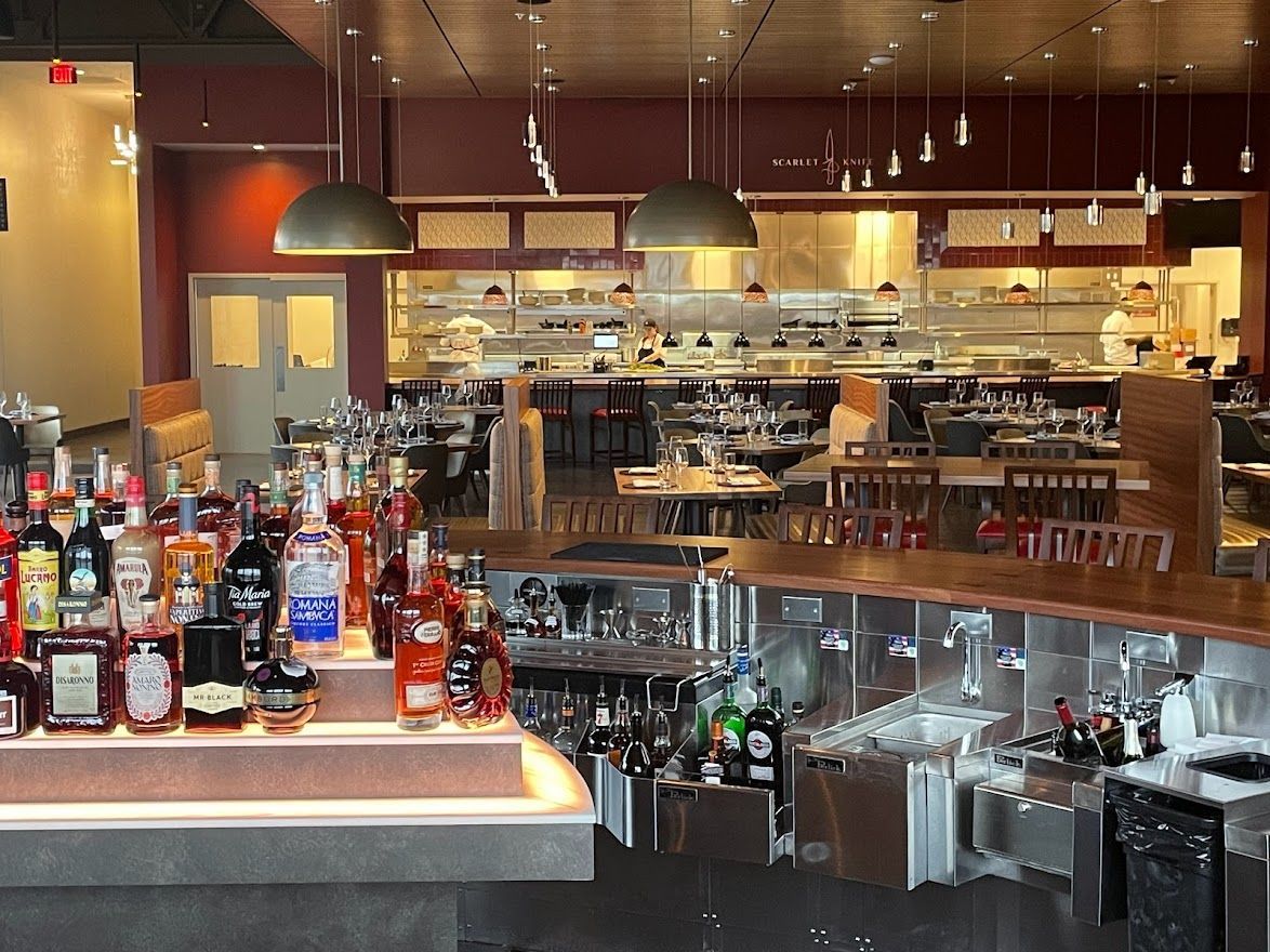 A bar with a tiered bottle display in the foreground and a dining room with tables and a kitchen area in the background.