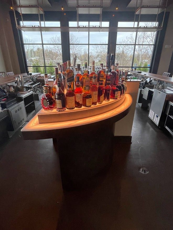 A curved wooden bar display holding a collection of liquor bottles in a room with large windows overlooking trees.