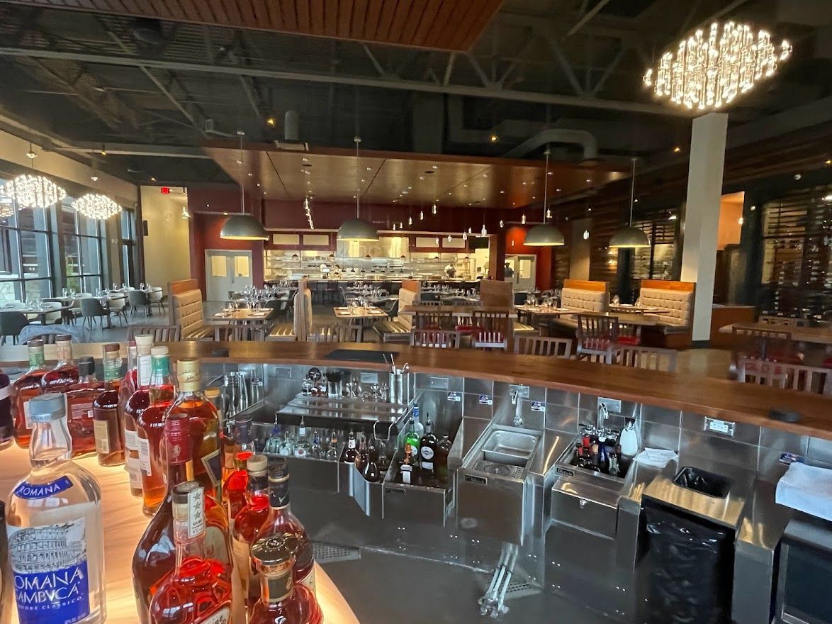A view from behind a bar top, showing liquor bottles in the foreground and a spacious, dimly lit restaurant dining area.