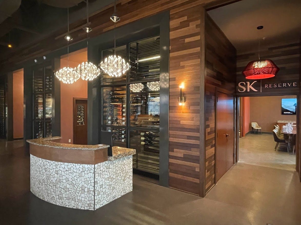 A reception area with a stone-fronted desk, decorative hanging lights, wood-paneled walls, and a wine cellar display.