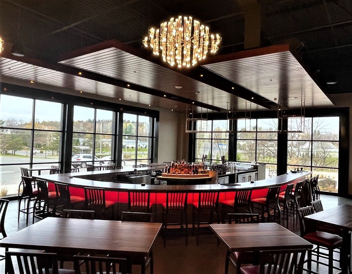 A curved restaurant bar with red-lit seating, wooden tables, and a modern tiered chandelier, framed by large glass walls.