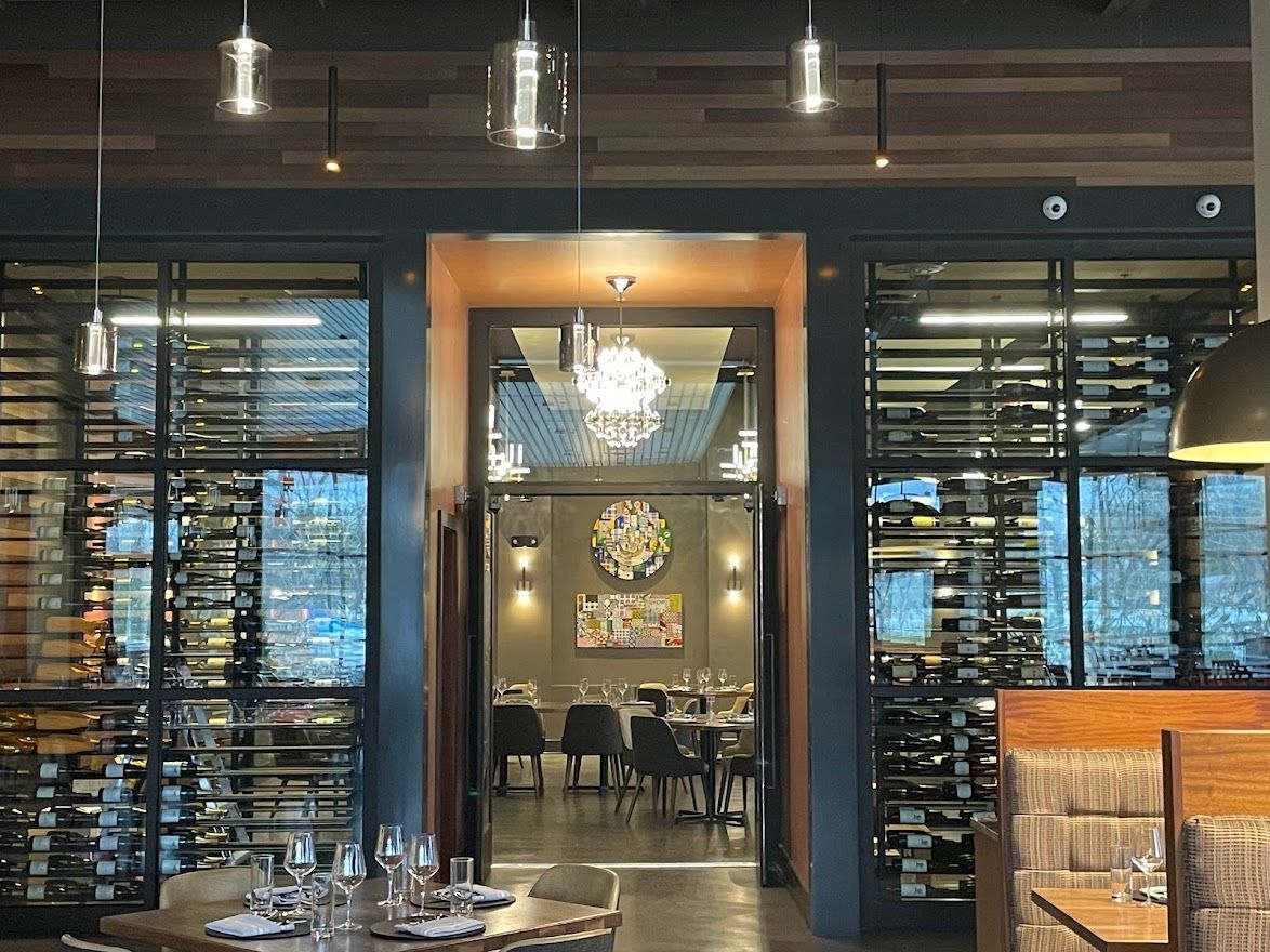 A dimly lit restaurant interior features large wine racks, warm overhead lights, and a view into a private dining area.
