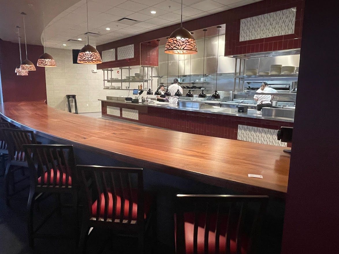 A long wooden bar counter with high chairs in front of an open kitchen with chefs working in a dimly lit restaurant.