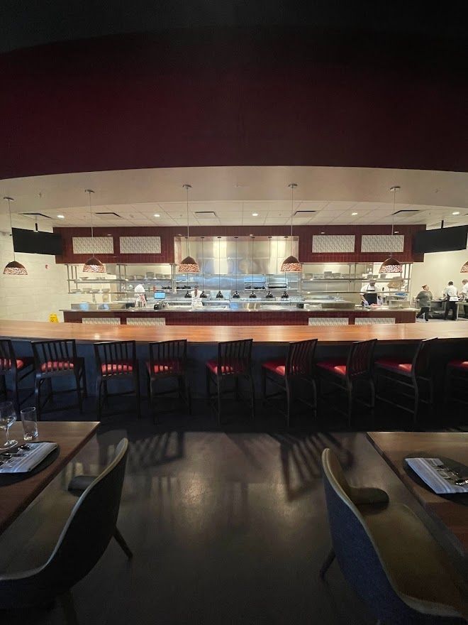 A restaurant interior features a long wooden bar with high stools facing a stainless-steel open kitchen under warm lights.