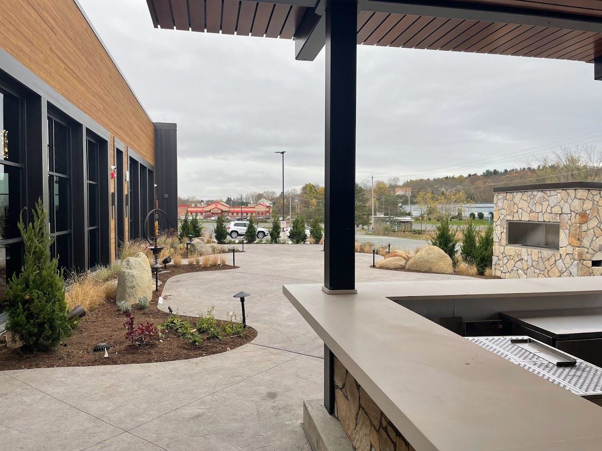 A modern outdoor patio with a stone bar and fire feature, adjacent to a commercial building with large glass windows.