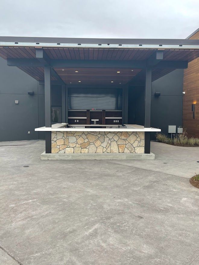 An outdoor bar with a stone base, a white countertop, and a wooden-paneled roof, set against a dark gray building.