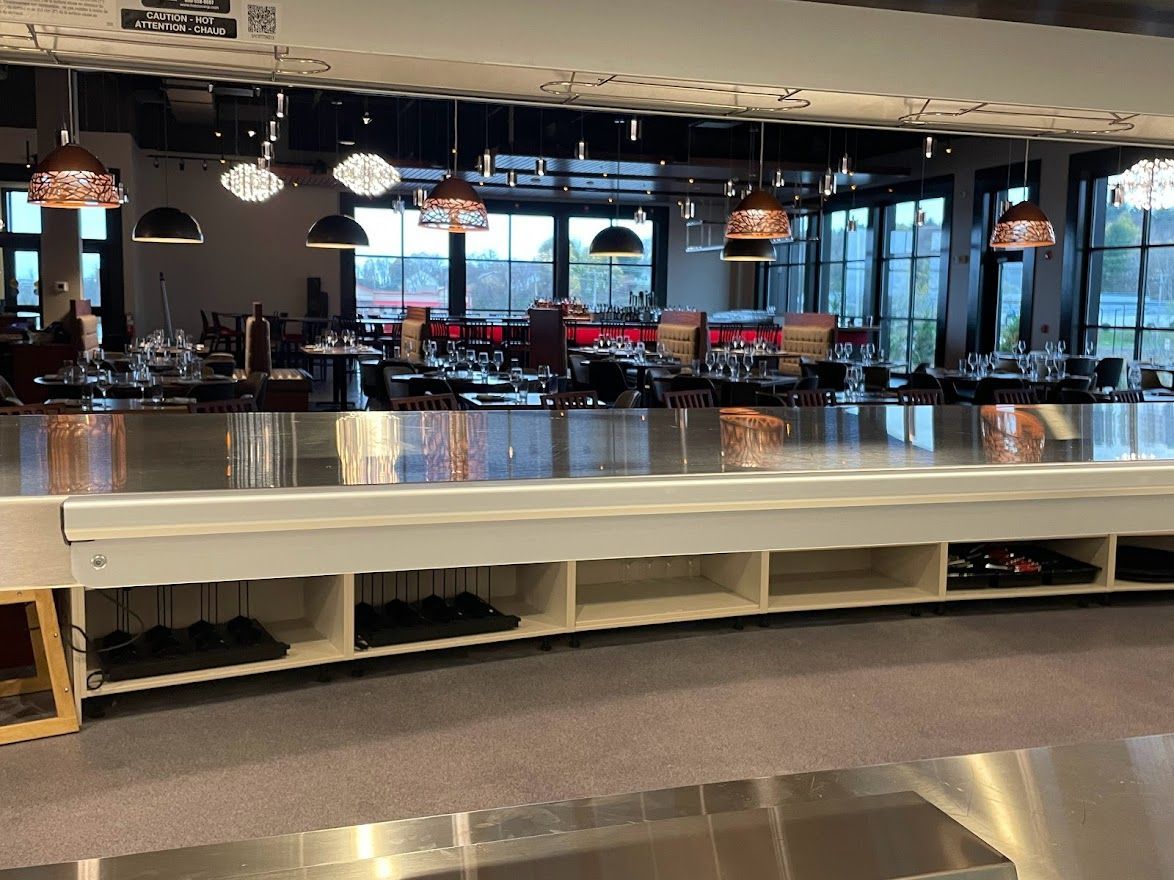 A restaurant bar counter with built-in lower shelves for glassware, set in a dining room with pendant lighting.