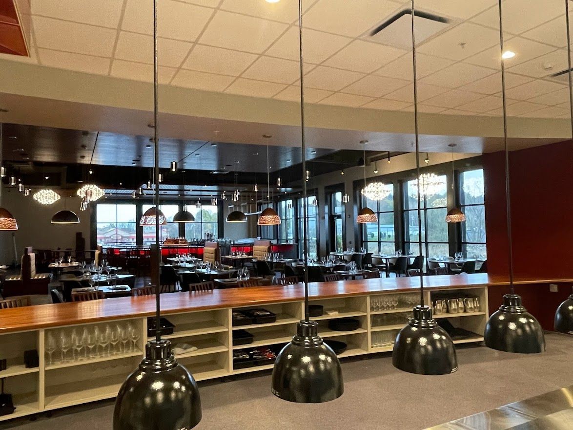 A wooden bar counter with white shelves sits in front of a dining room with tables, chairs, and hanging pendant lights.