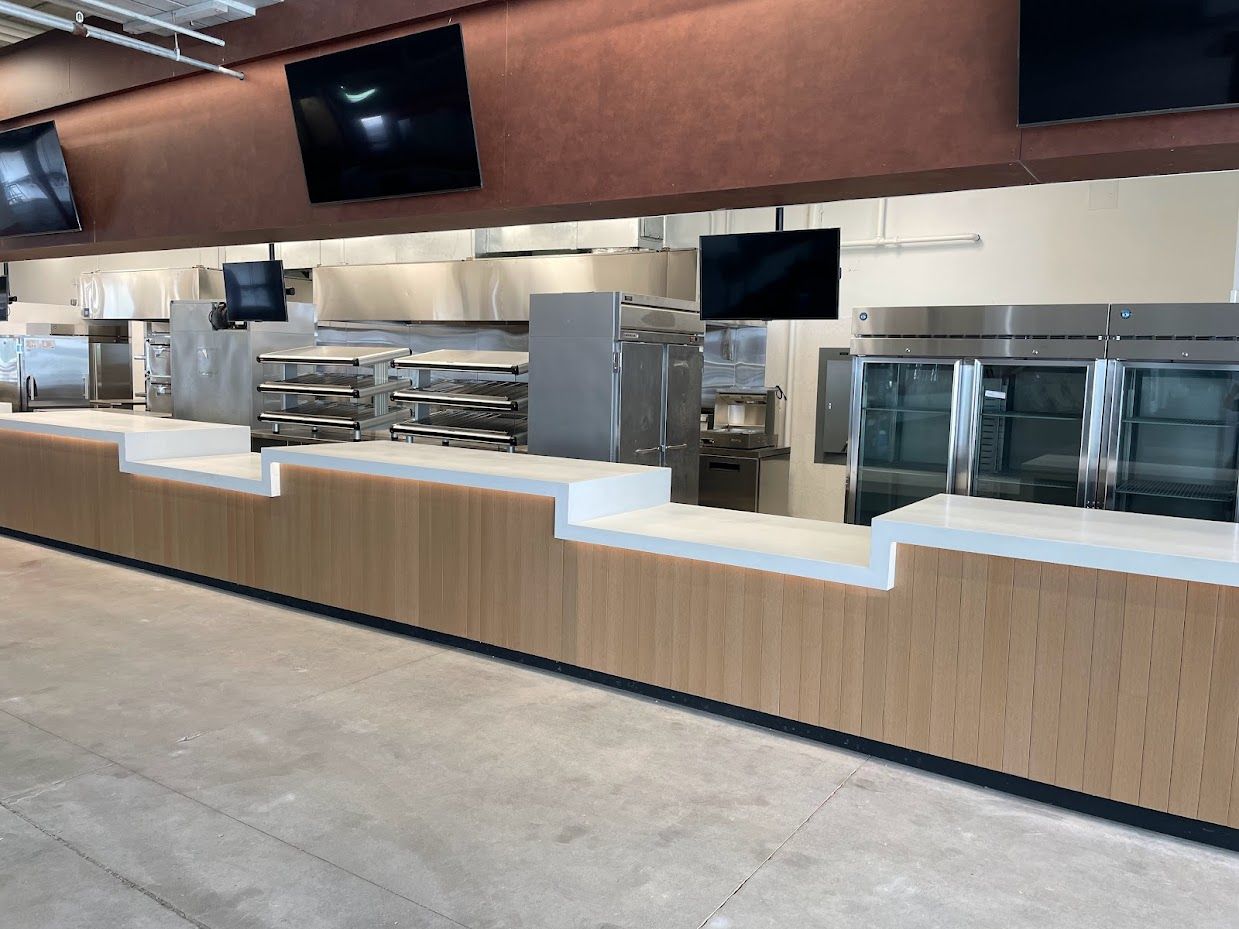 A modern serving counter in a kitchen with white surfaces, light wood-paneled walls, and black digital menu screens.