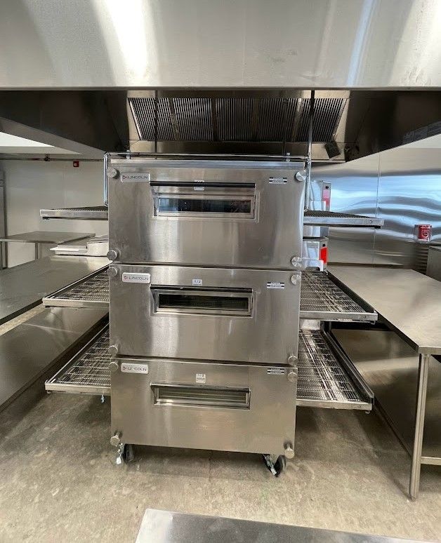 A stainless steel, three-deck commercial pizza oven on wheels situated under a vent hood in a professional kitchen.