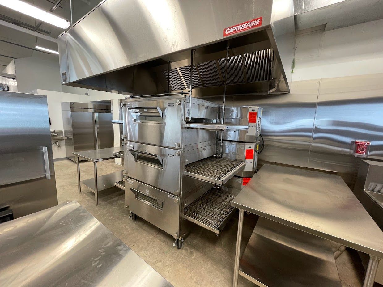 Industrial commercial kitchen with a stacked pizza oven under a large stainless steel exhaust hood and metal tables.