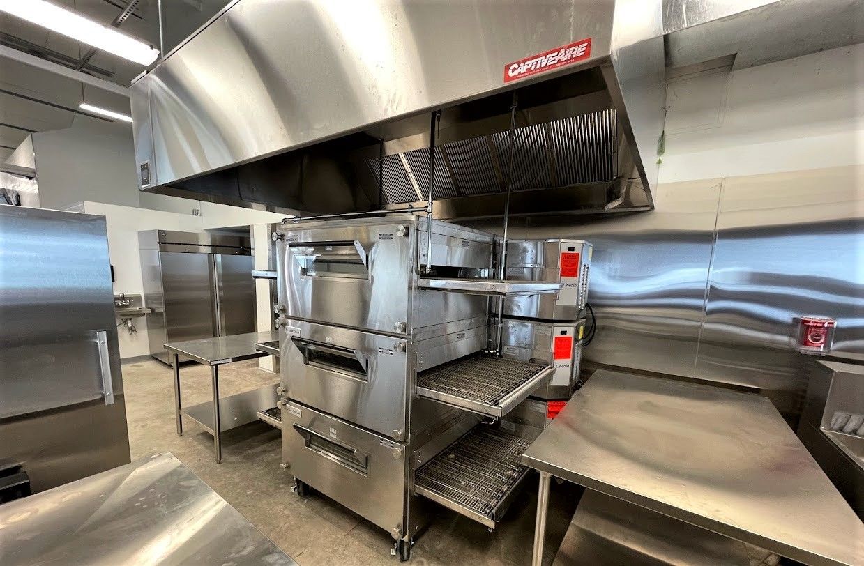 A multi-deck commercial pizza oven positioned under a stainless steel vent hood in a professional kitchen.