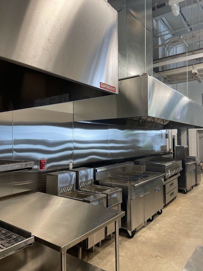 A professional commercial kitchen featuring stainless steel prep tables, cooking ranges, and large exhaust hoods.