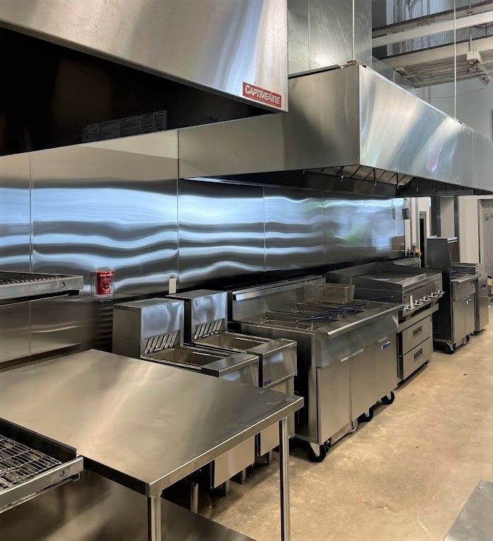 A commercial kitchen featuring stainless steel prep tables, ranges, and large exhaust hoods over a tiled floor.