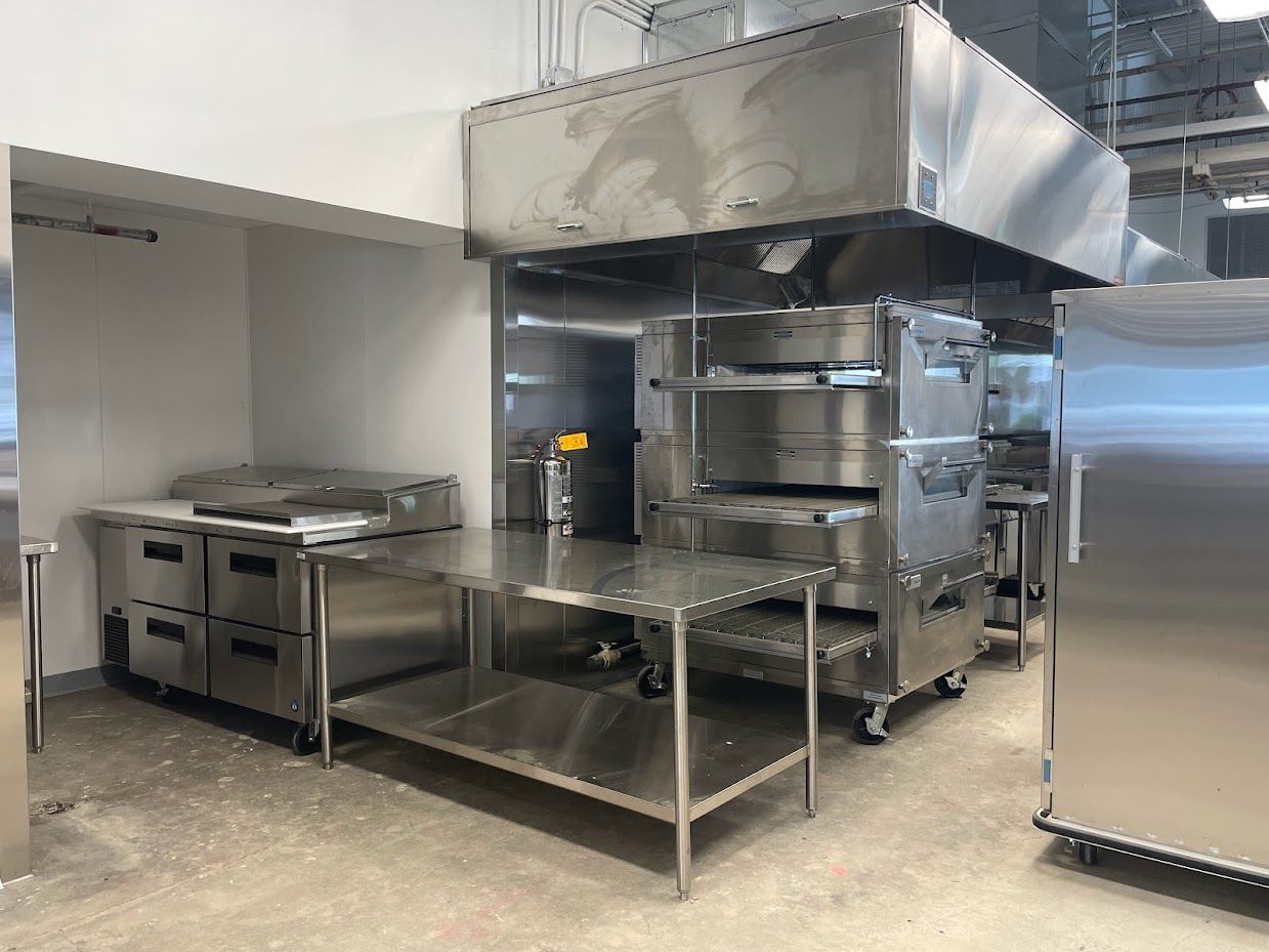 A stainless steel commercial kitchen featuring a multi-deck oven under a large vent hood, a prep table, and work stations.
