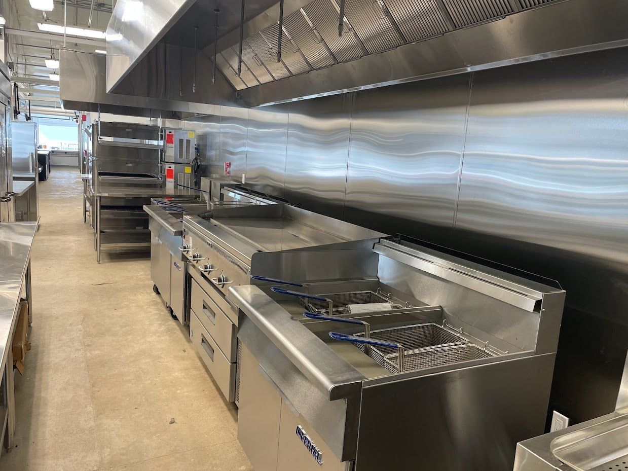 A clean, professional stainless steel commercial kitchen line with fryers, a flat-top grill, and overhead vent hoods.