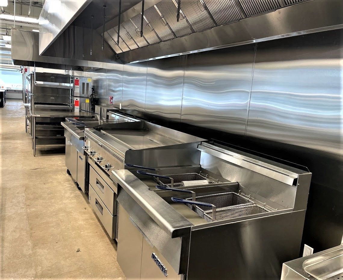 A professional stainless steel kitchen line featuring fryers, a flat-top grill, and an overhead ventilation hood.