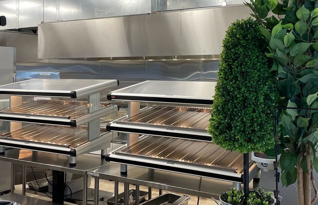 Two stainless steel, multi-tiered kitchen warming racks sit indoors next to a decorative topiary plant.