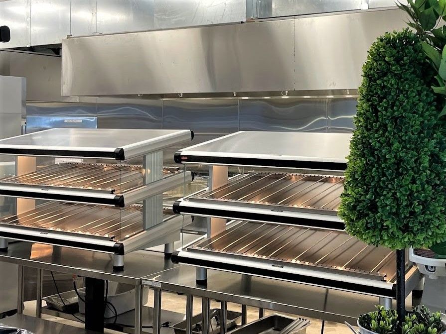 Two stainless steel multi-tier food warming stations in a commercial kitchen next to a decorative topiary tree.