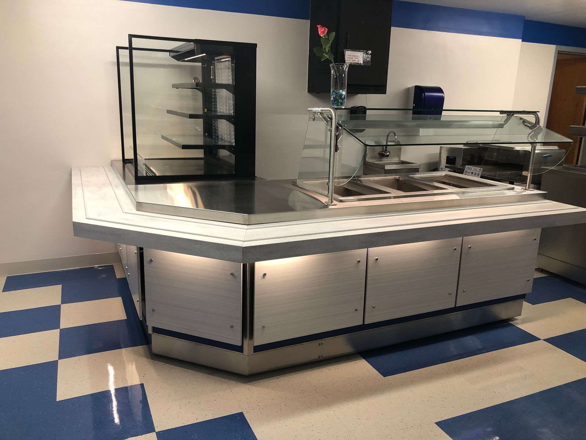 A buffet serving counter with a food warmer, sneeze guard, and shelf unit set on a blue-and-white checkered floor.
