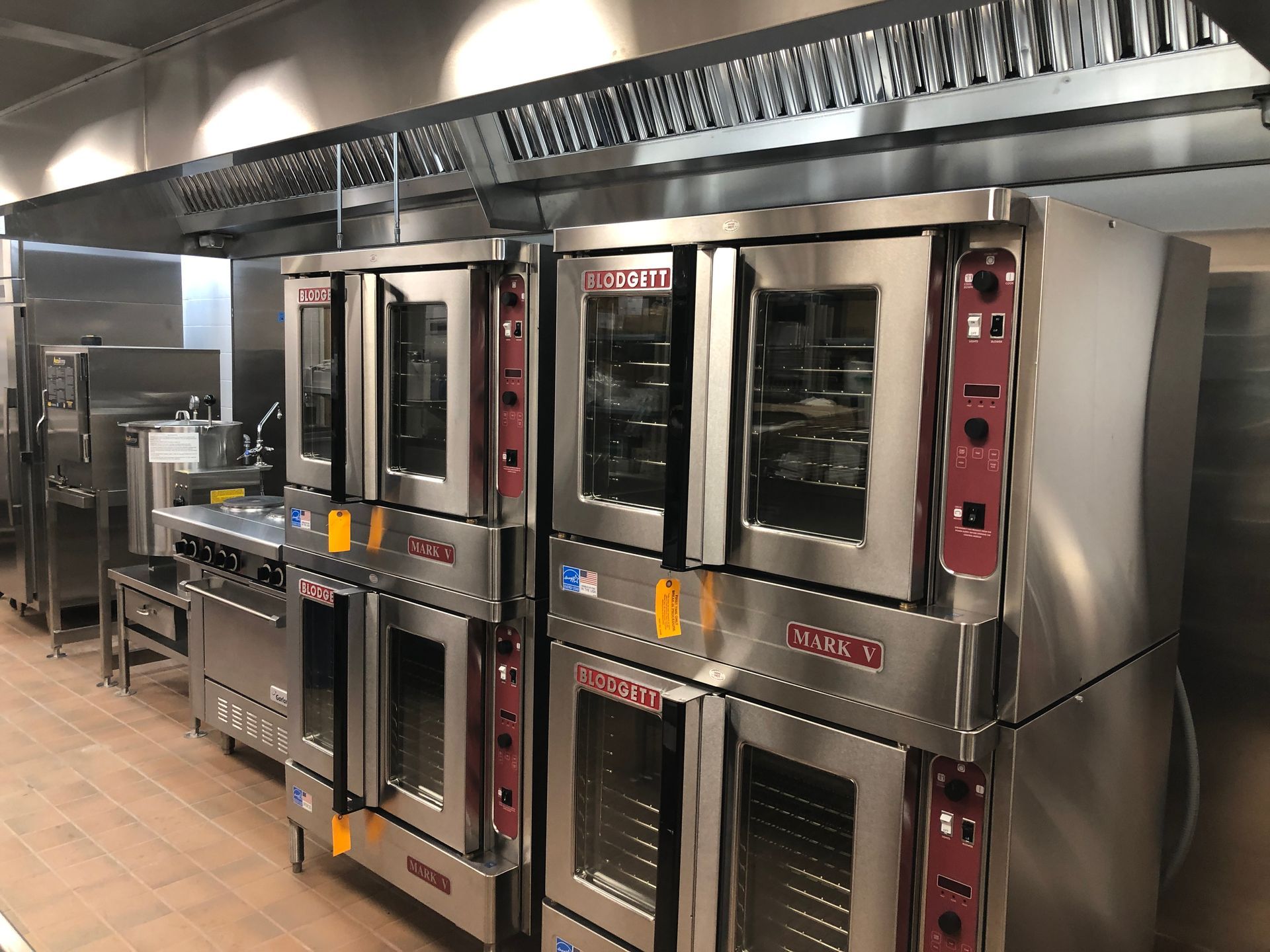 A row of stainless steel commercial ovens stands in a professional kitchen under a metallic ventilation hood.