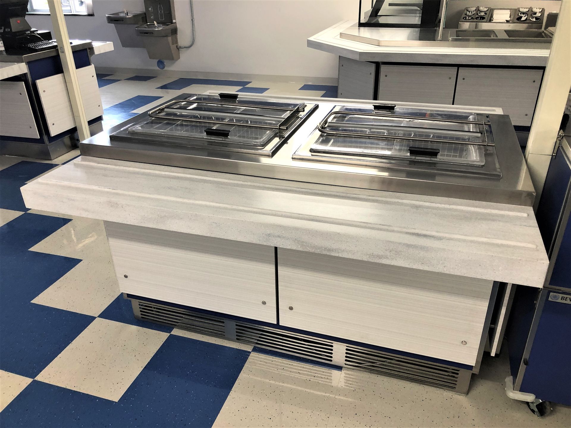 A cafeteria food serving station with two covered stainless steel food wells, white cabinets, and a checkered floor.
