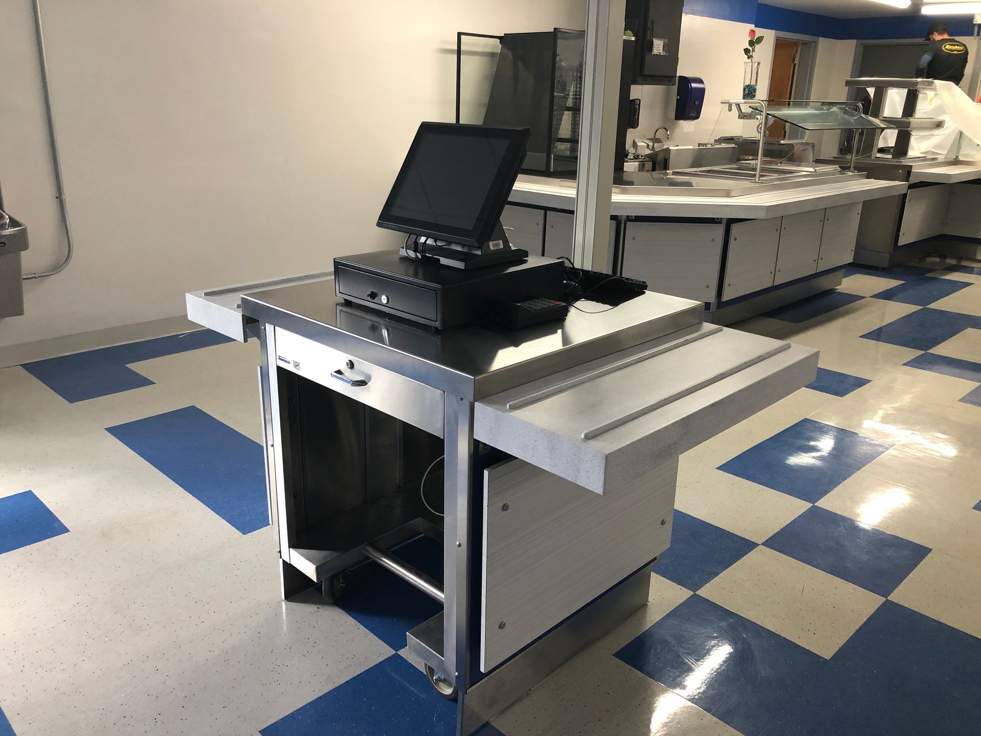 A point-of-sale terminal on a stainless steel stand in a room with a blue and white checkered floor.