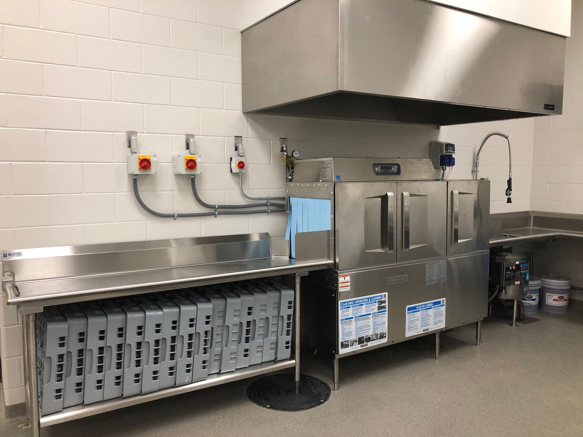 A commercial kitchen dishwashing station featuring a stainless steel hood, machine, and prep table with stacked crates.