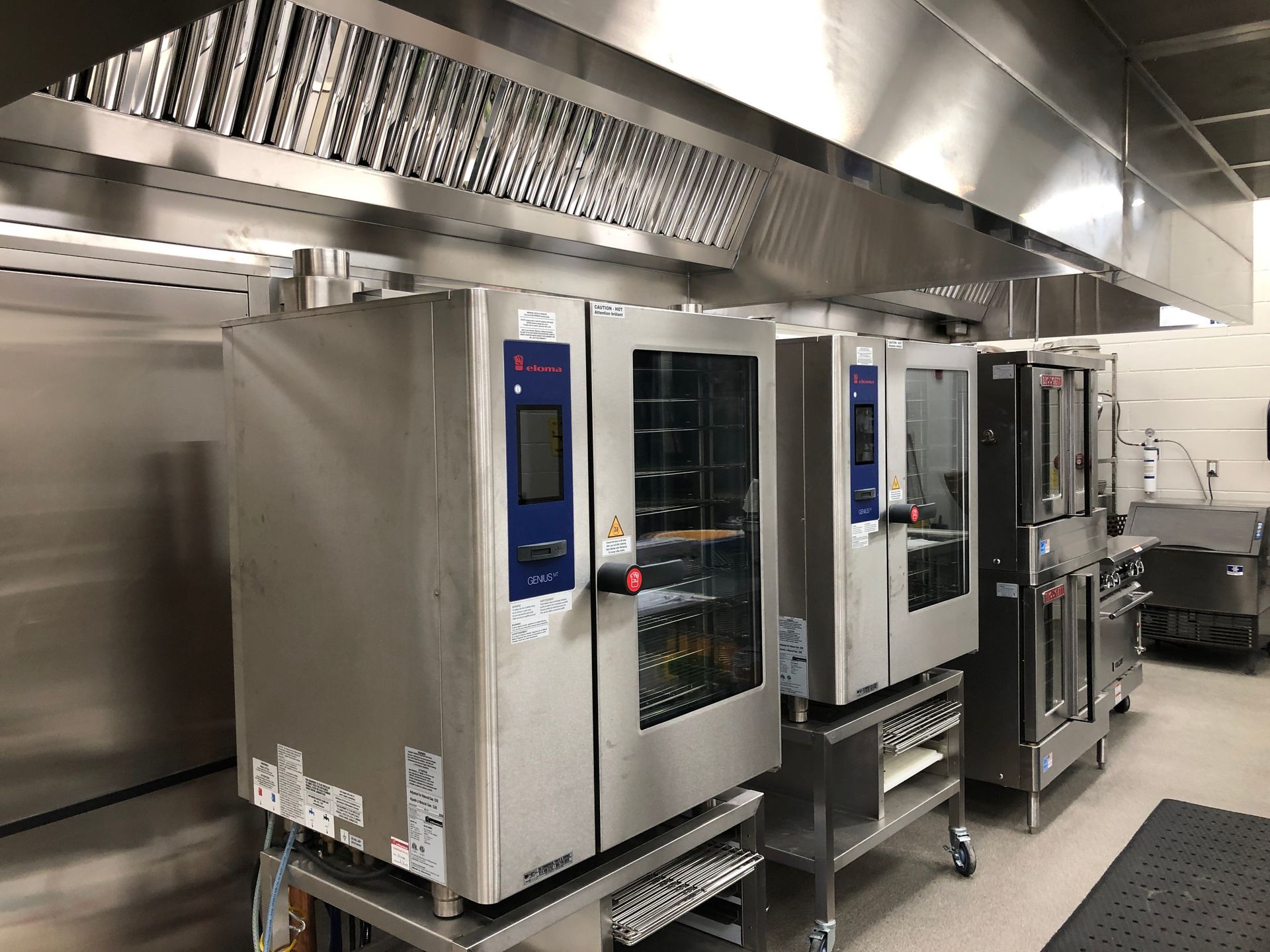 A row of stainless steel commercial combi ovens sits under a large metal ventilation hood in a kitchen.