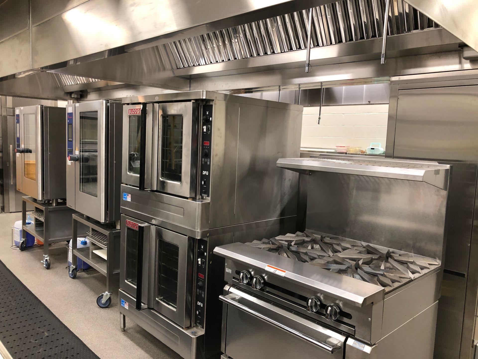 A row of stainless steel commercial kitchen appliances, including stacked ovens and a gas range under a metal hood.