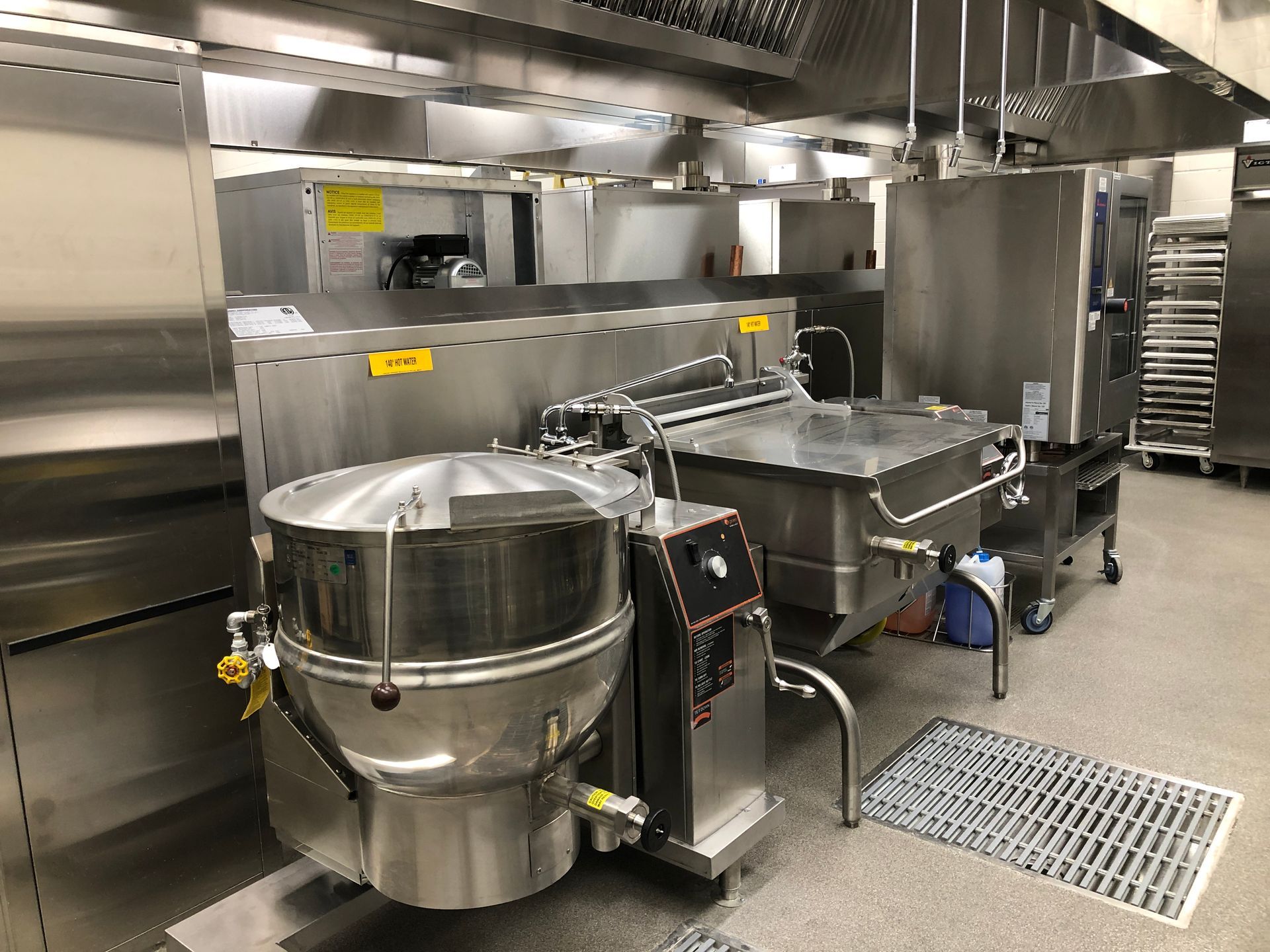 A commercial kitchen featuring stainless steel equipment, including a tilt skillet and a steam kettle under a vent hood.