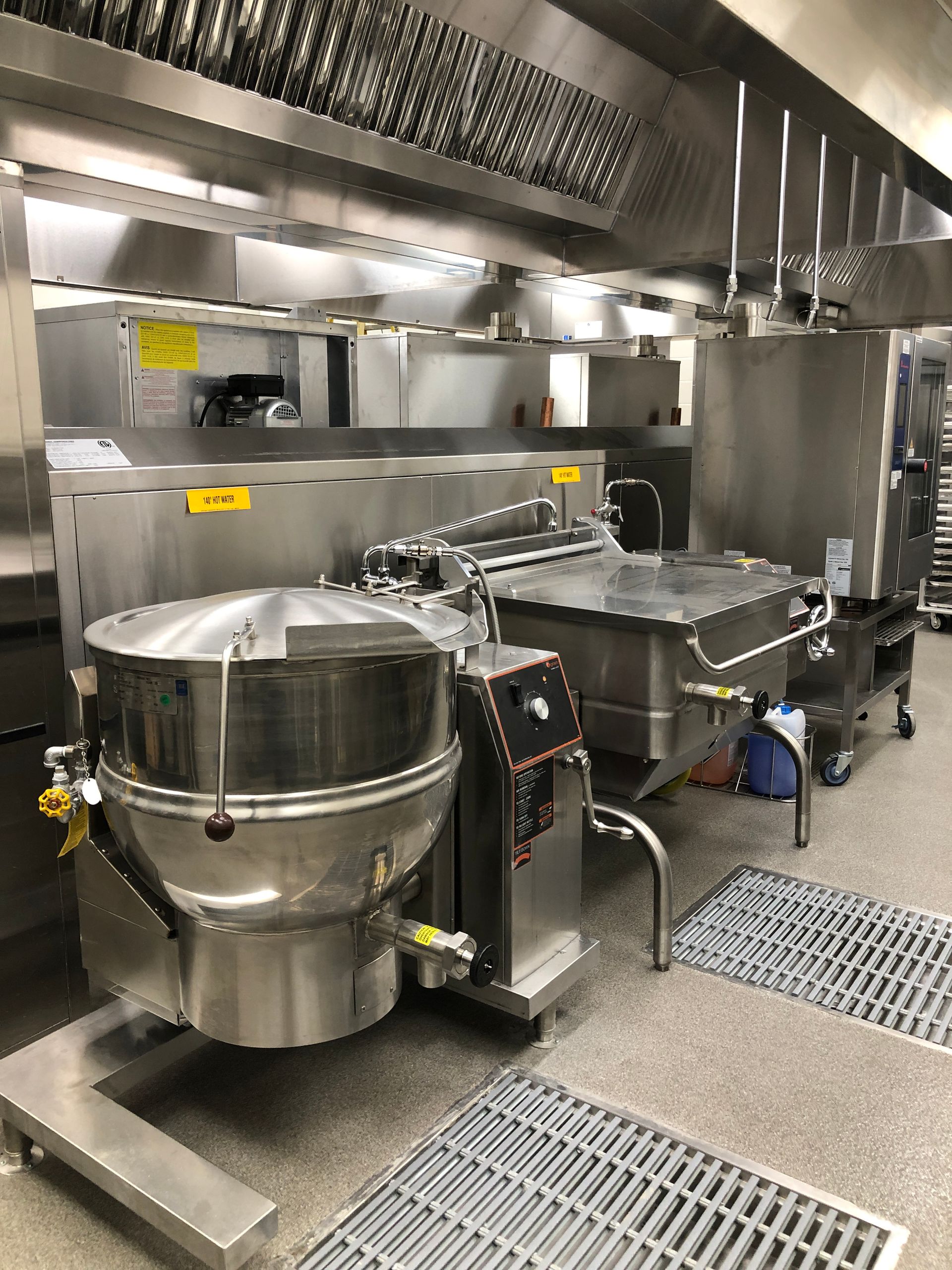 Professional industrial kitchen with large stainless steel tilting skillets and a soup kettle under a ventilation hood.