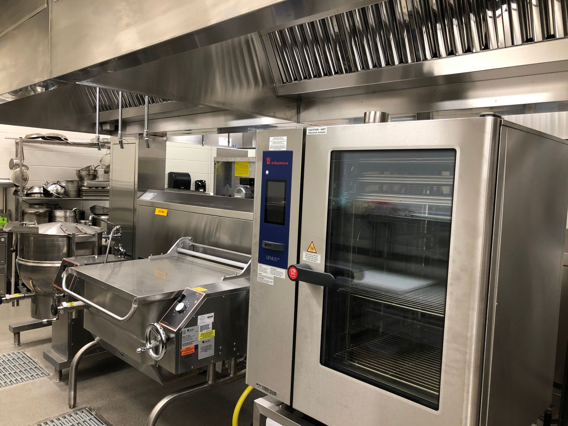 A professional kitchen featuring a large stainless steel industrial oven and a tilt skillet under a ventilation hood.