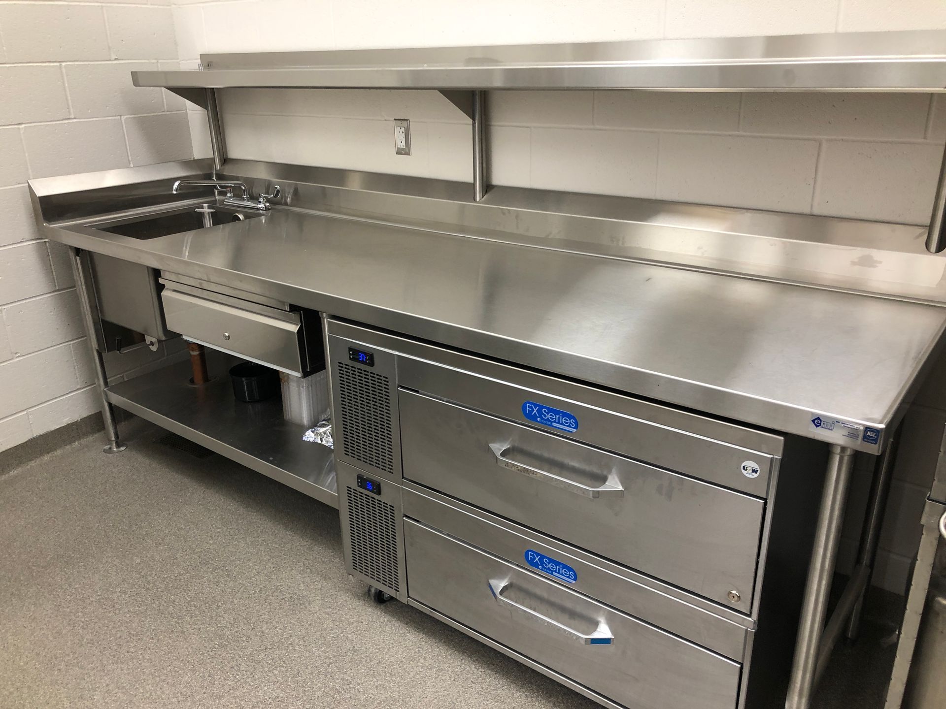 Stainless steel commercial kitchen workstation with a sink, prep counter, shelving, and two built-in refrigerated drawers.