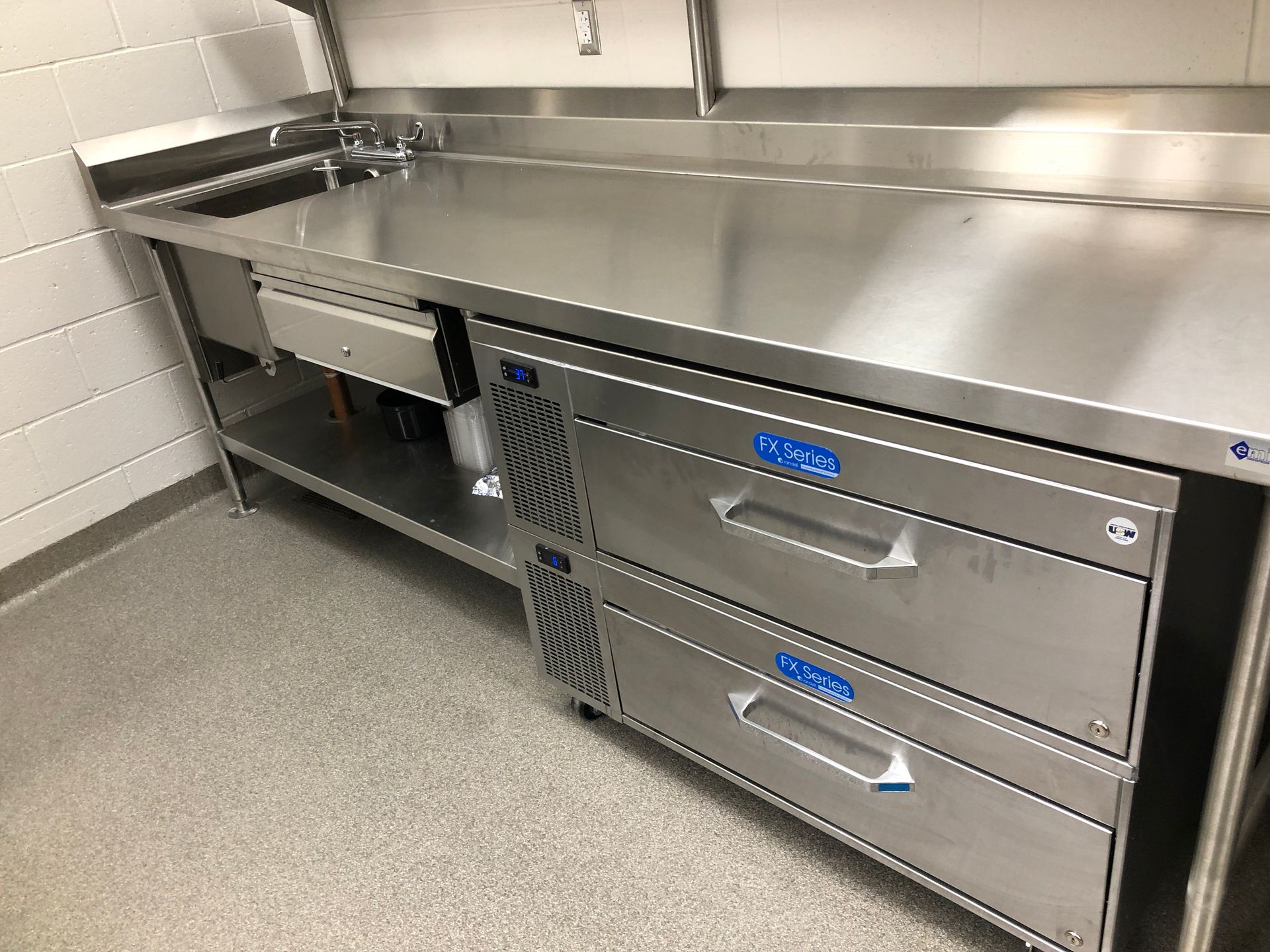 A stainless steel work table with a built-in, two-drawer under-counter refrigerator in a commercial kitchen setting.