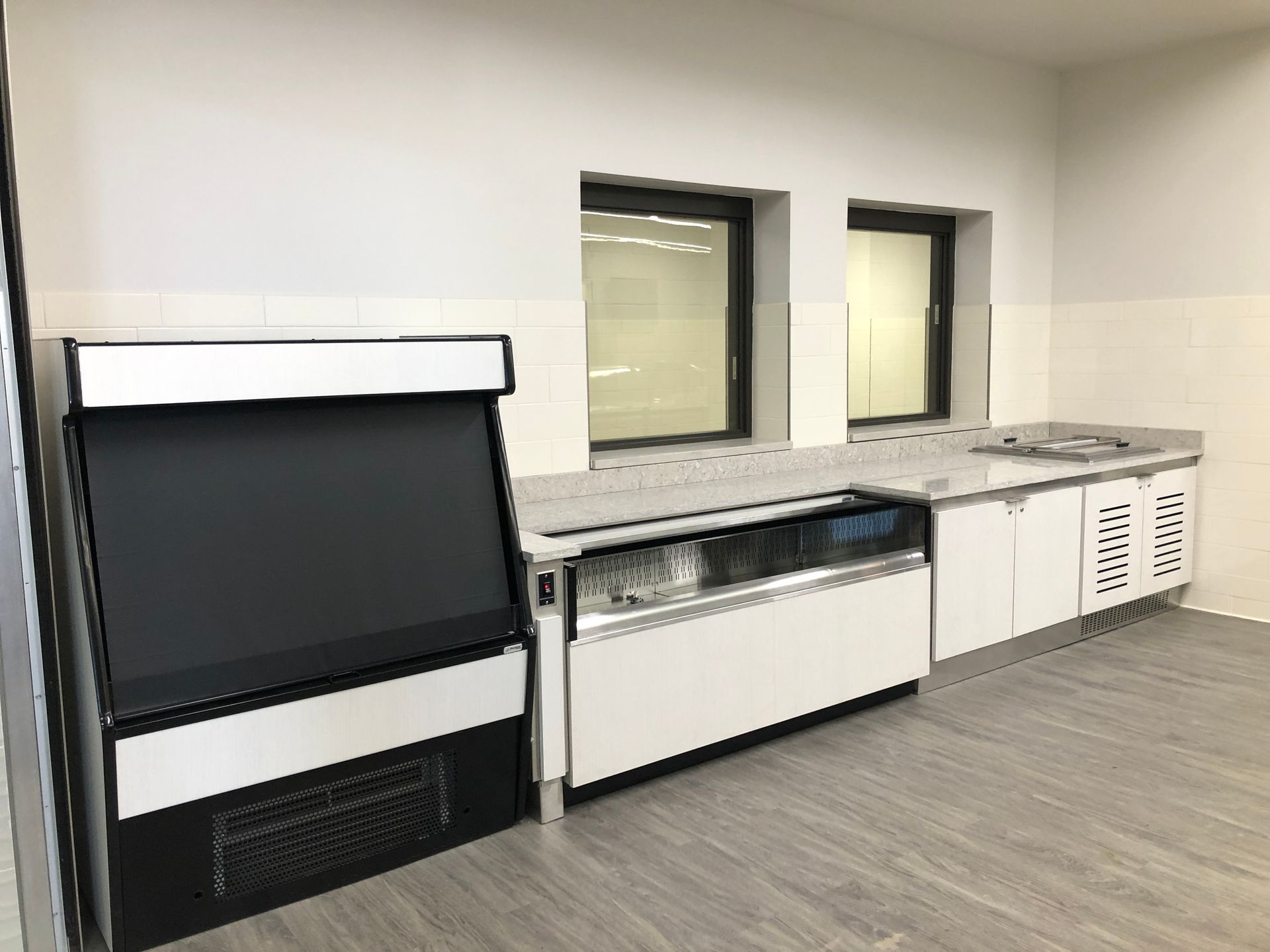 A black upright refrigerator stands next to a low, white refrigerated display counter with cabinets against a white wall.