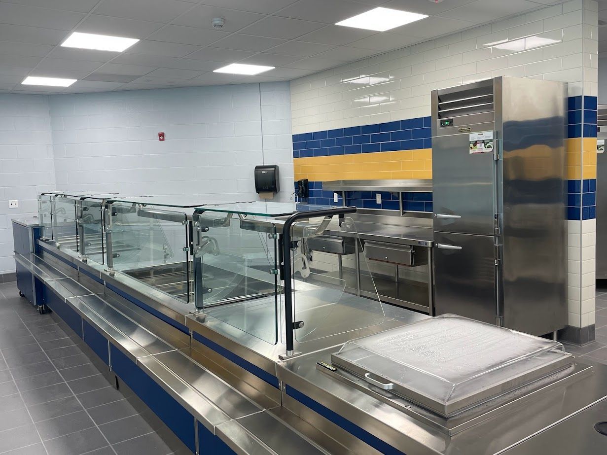 A stainless steel cafeteria serving counter with a glass sneeze guard, located in a room with blue and yellow wall tiles.