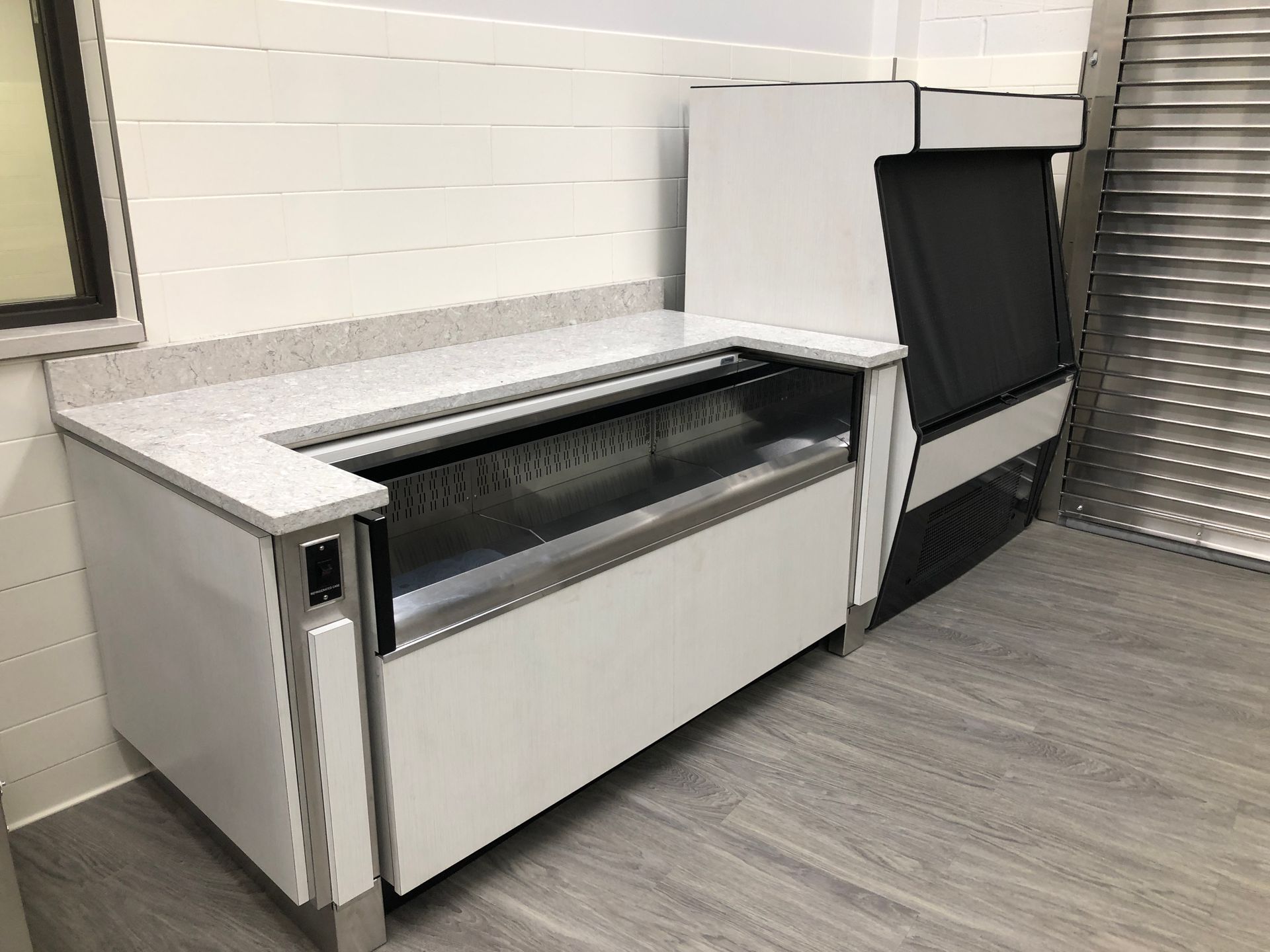 White and silver commercial refrigeration unit with a countertop and adjacent display case on a grey wood-look floor.