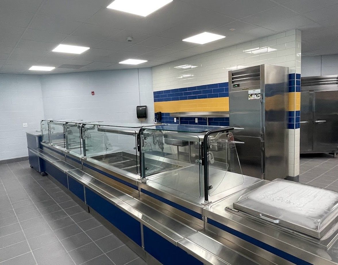A modern cafeteria serving line with stainless steel equipment, glass food guards, and blue and yellow tiled walls.
