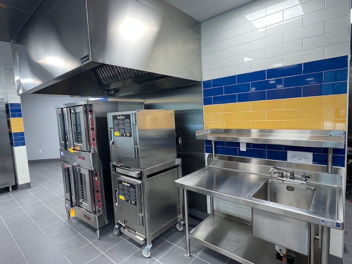 Commercial kitchen area with stainless steel ovens, a vent hood, and a metal prep table with a sink against tiled walls.