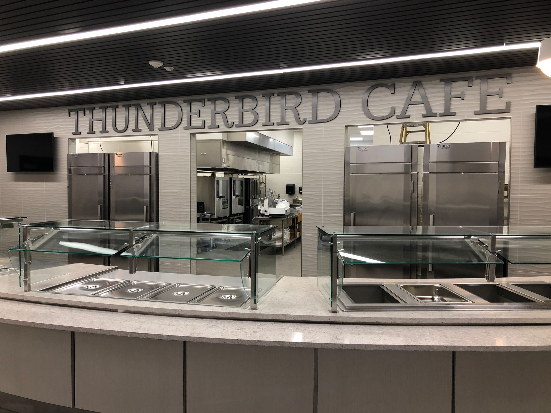 A commercial cafeteria serving line with a marble countertop, glass sneeze guards, and a kitchen visible behind it.