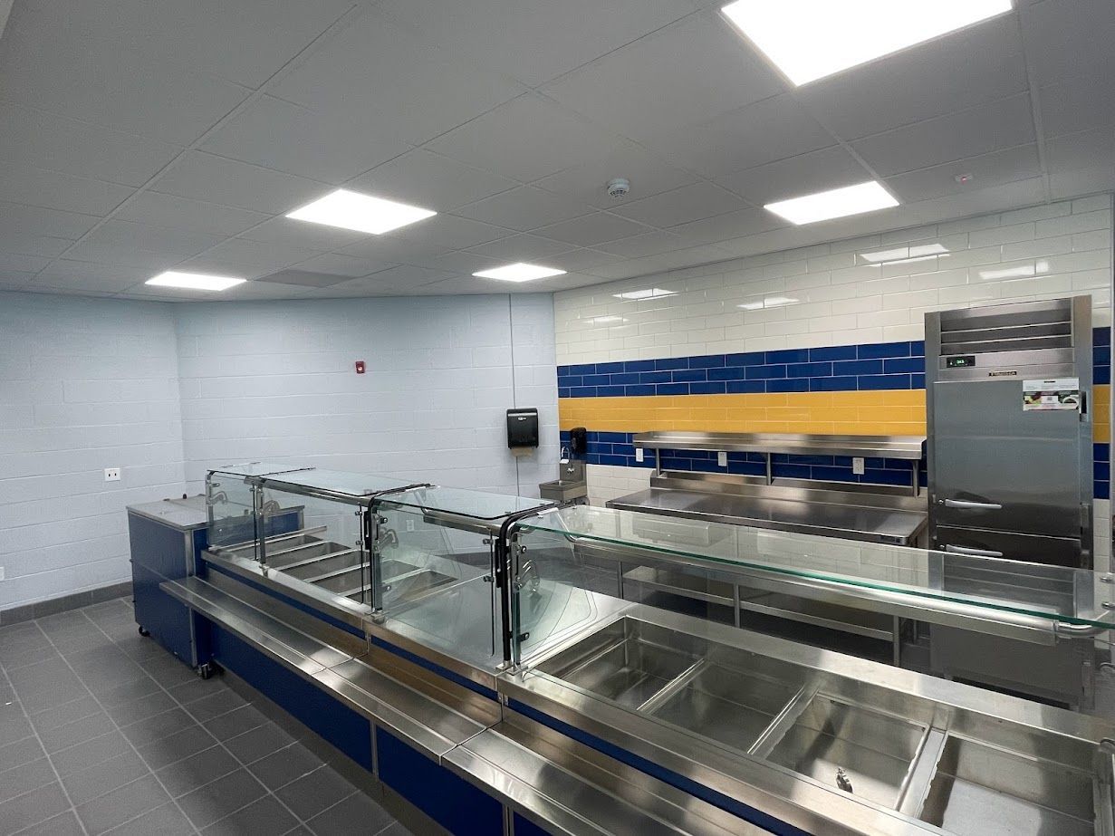 A cafeteria food service counter featuring stainless steel food wells and a glass sneeze guard against a blue-tiled wall.