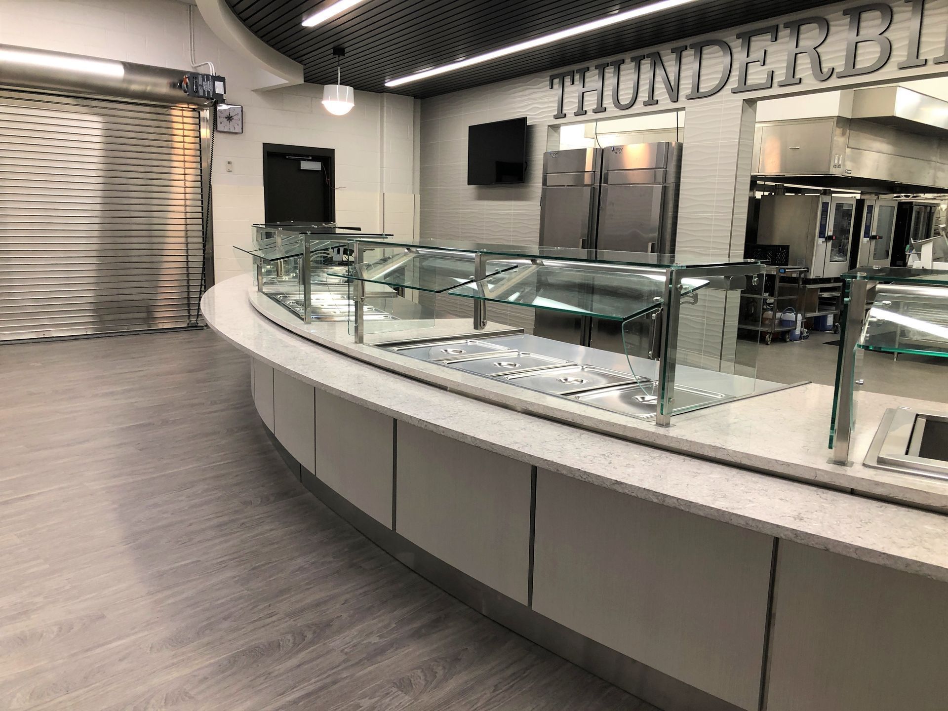 A modern, curved serving counter with a glass sneeze guard, light gray cabinetry, and wooden flooring in a cafe setting.