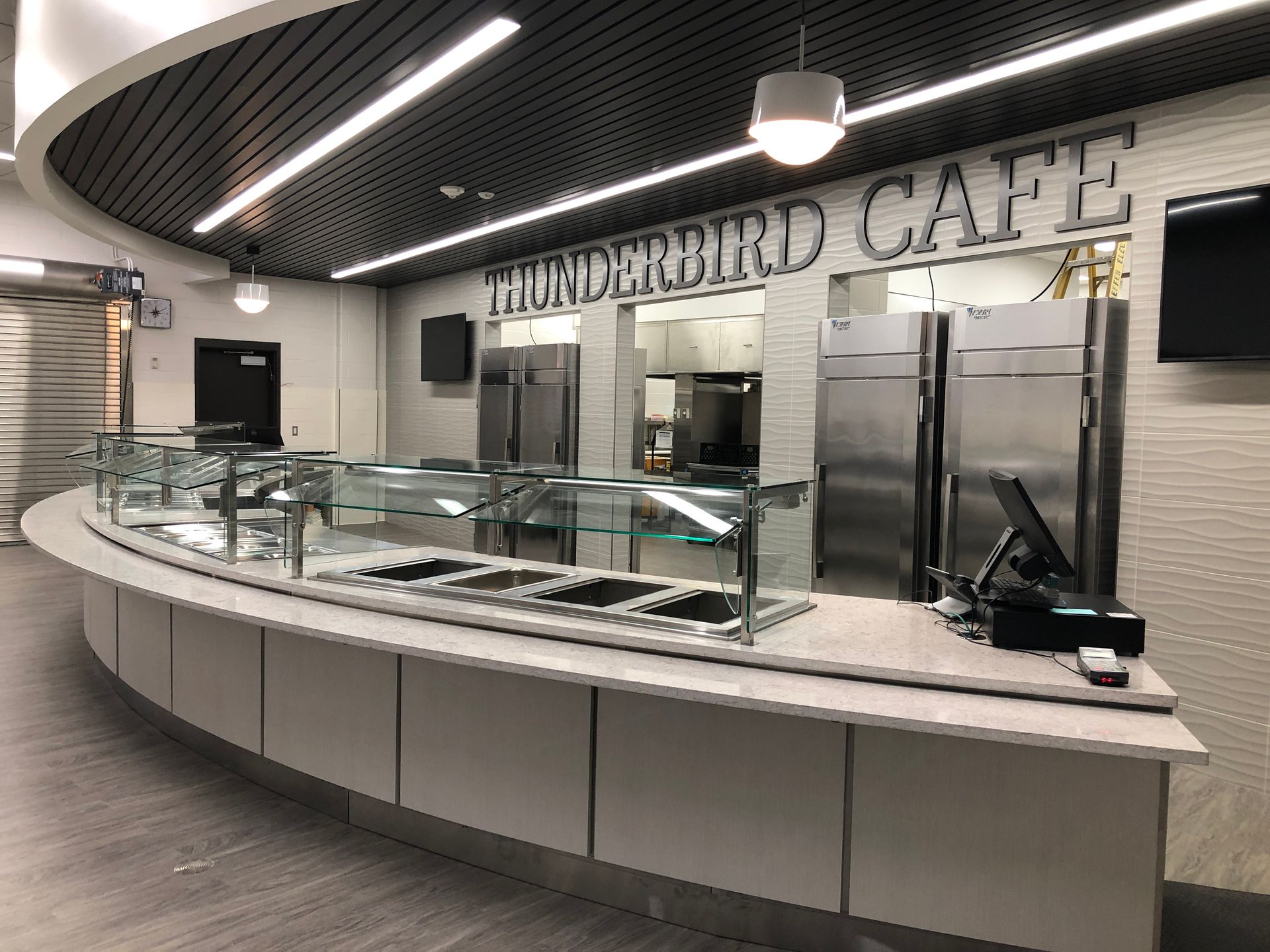 A modern, curved serving counter at the Thunderbird Cafe, featuring stainless steel food pans, refrigerators, and lighting.